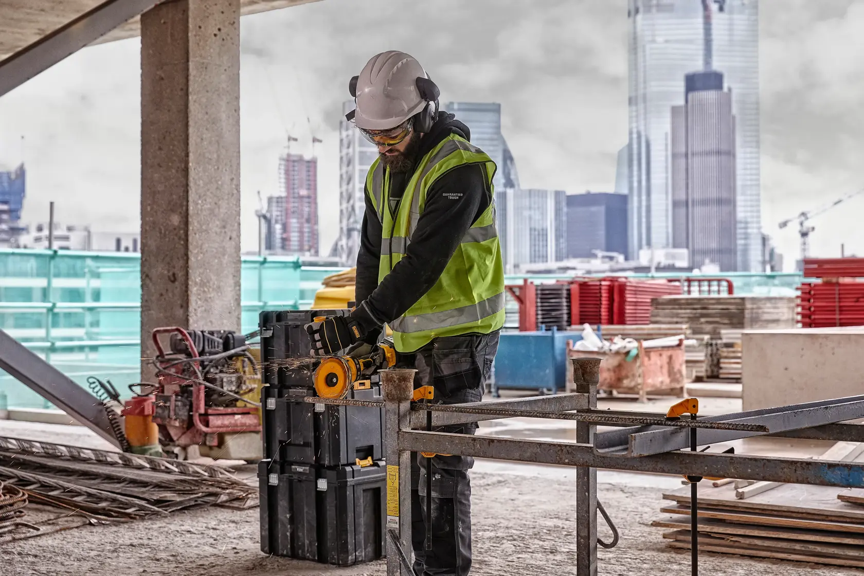 Worker uses DEWALT DCG418NT tool on construction site, wearing safety gear and surrounded by equipment and city buildings.