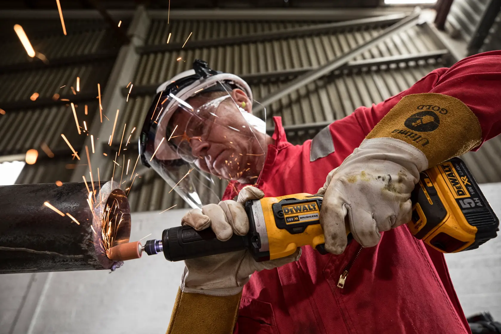 A person wearing protective gloves and safety gear uses a DEWALT DCG426N cordless grinder on a metal pipe, creating sparks in an industrial environment.