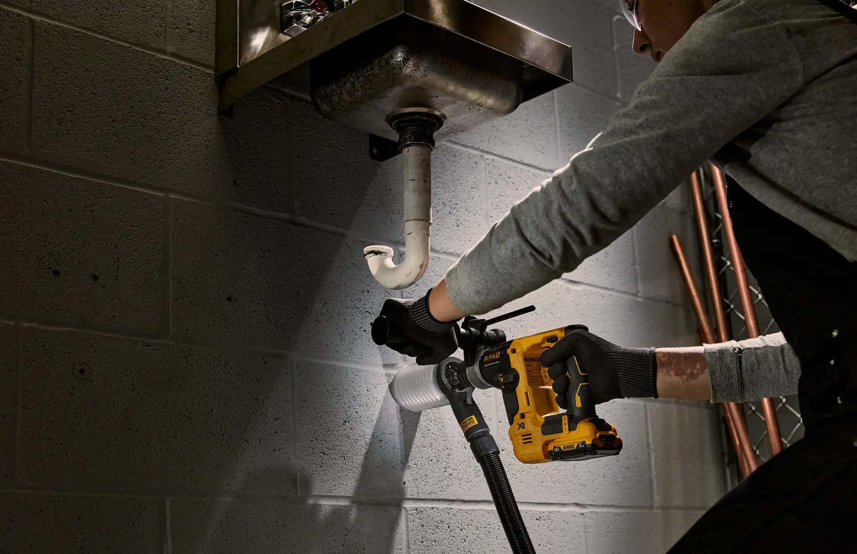 Person uses a DEWALT cordless drill under a sink to work on plumbing against a brick wall.