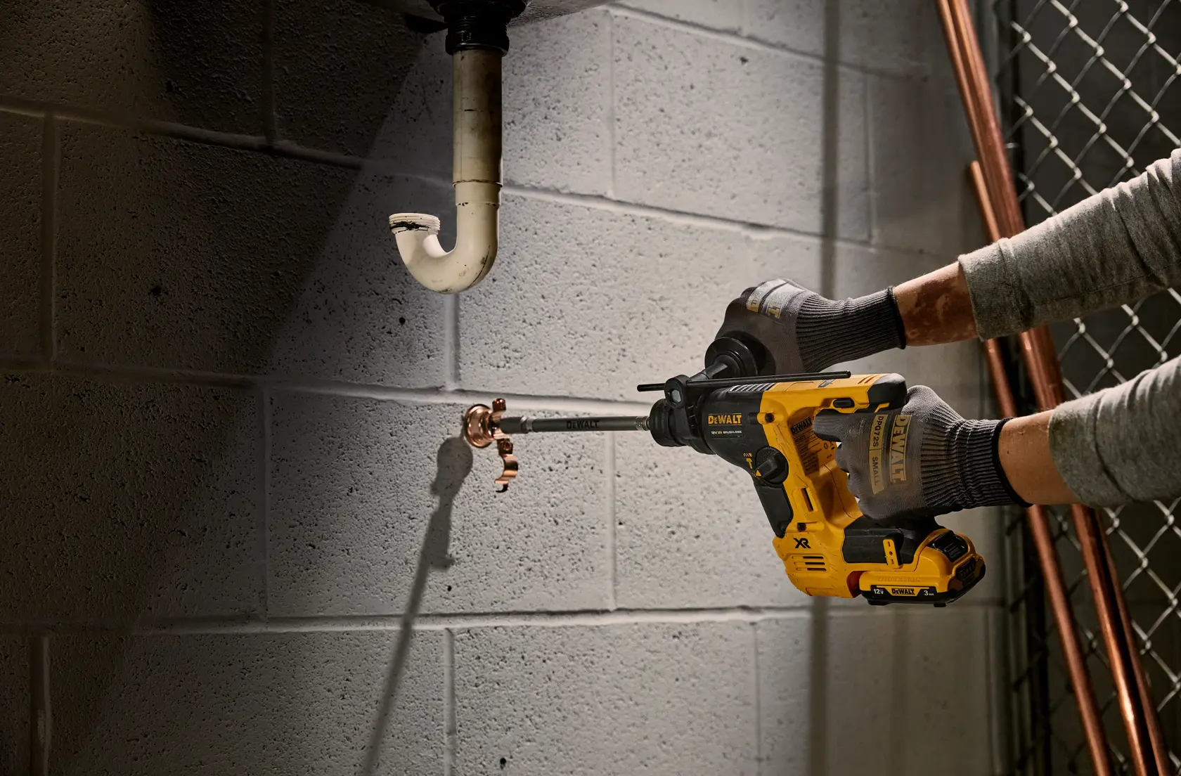 A person uses a DEWALT rotary hammer drill to bore into a concrete wall near plumbing pipes.