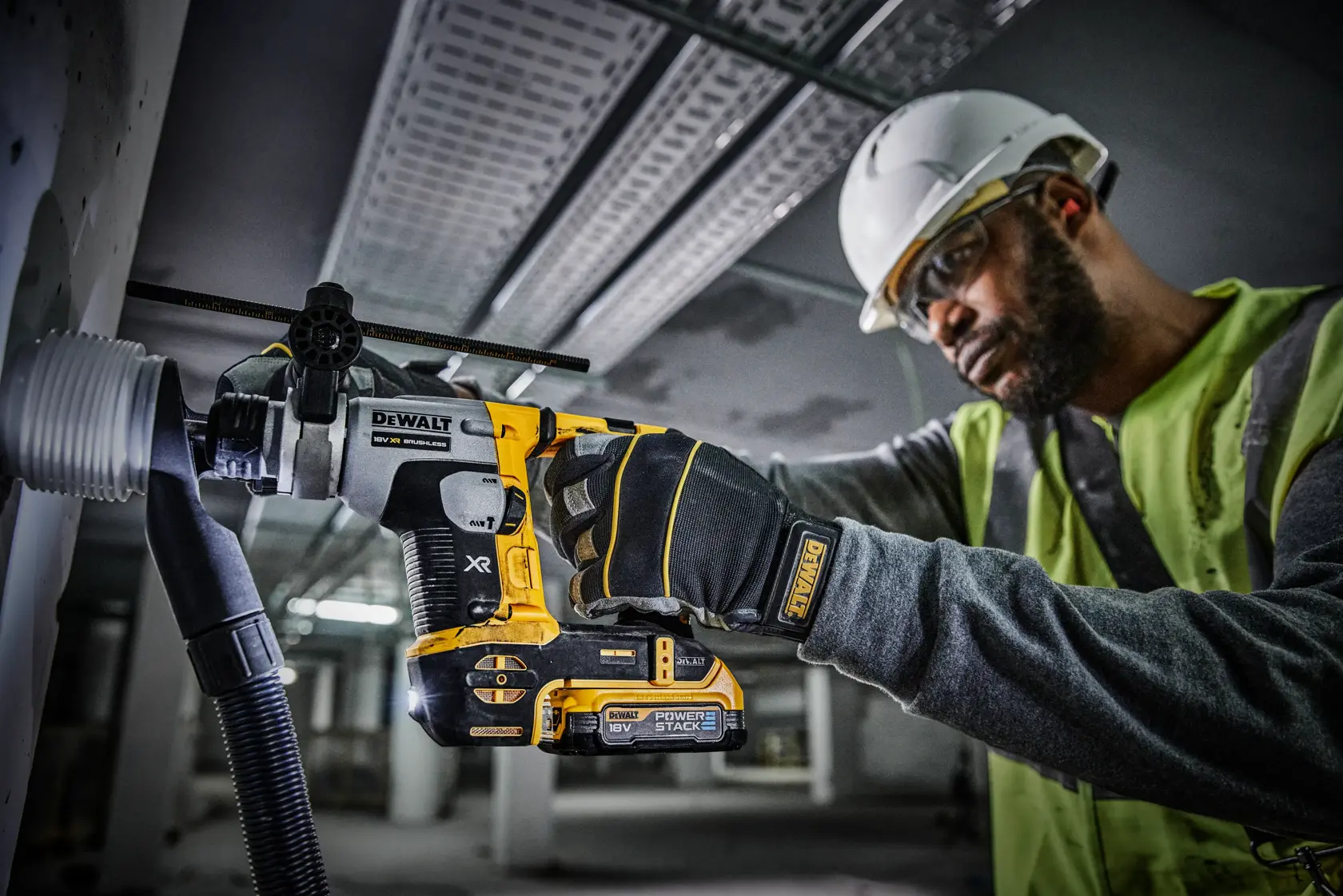 A person wearing a safety helmet and high-visibility vest is using a DEWALT cordless rotary hammer drill to bore into a wall, with dust extraction equipment attached. The drill is labeled with XR and Power Stack branding.