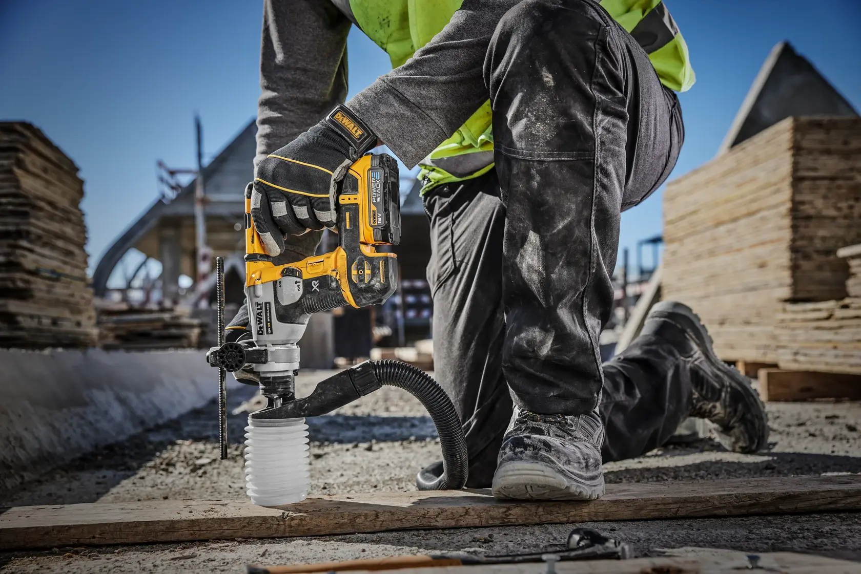 A construction worker is using a DEWALT cordless rotary hammer drill (model DCH172E2T) with dust extraction on a building site, drilling into the ground while kneeling on one knee.