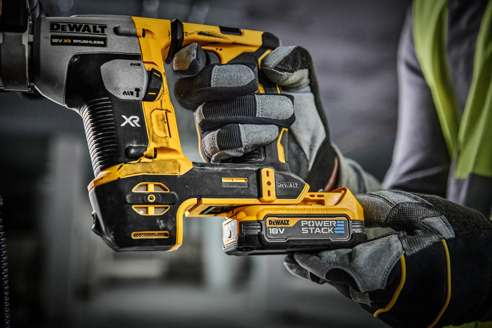 Close-up of a person wearing work gloves holding a DEWALT 18V XR brushless power tool with a Power Stack battery in a workshop or construction environment.