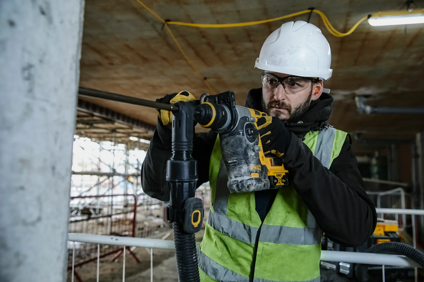 Worker using a DEWALT rotary hammer drill on a construction site, wearing a helmet, gloves, and safety vest.