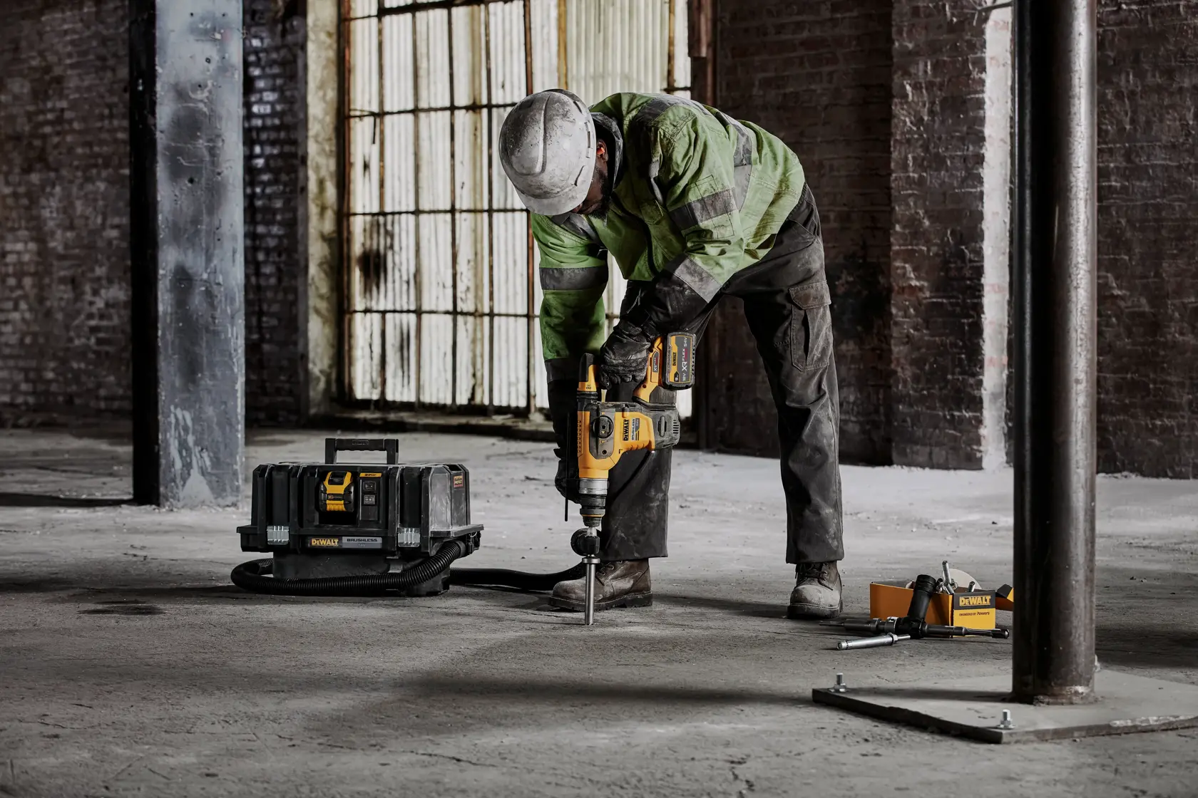 Person using DEWALT rotary hammer drill on concrete floor at a construction site with tool case nearby.