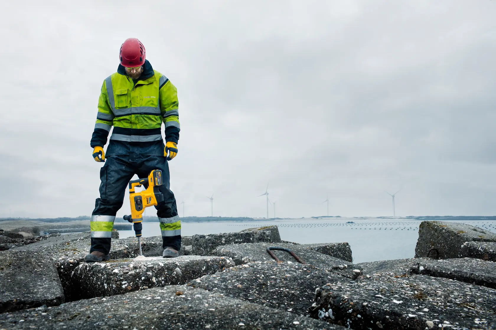 A person wearing high-visibility workwear and a red helmet is using a yellow DEWALT rotary hammer drill (SKU: DCH481N) to drill into concrete at an outdoor construction site by the water, with wind turbines visible in the background.