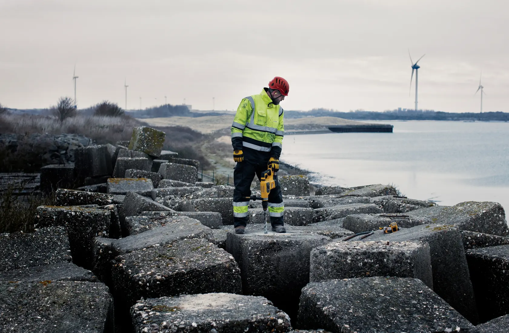 Worker in safety gear uses DEWALT tool on large concrete blocks near water with wind turbines in background.