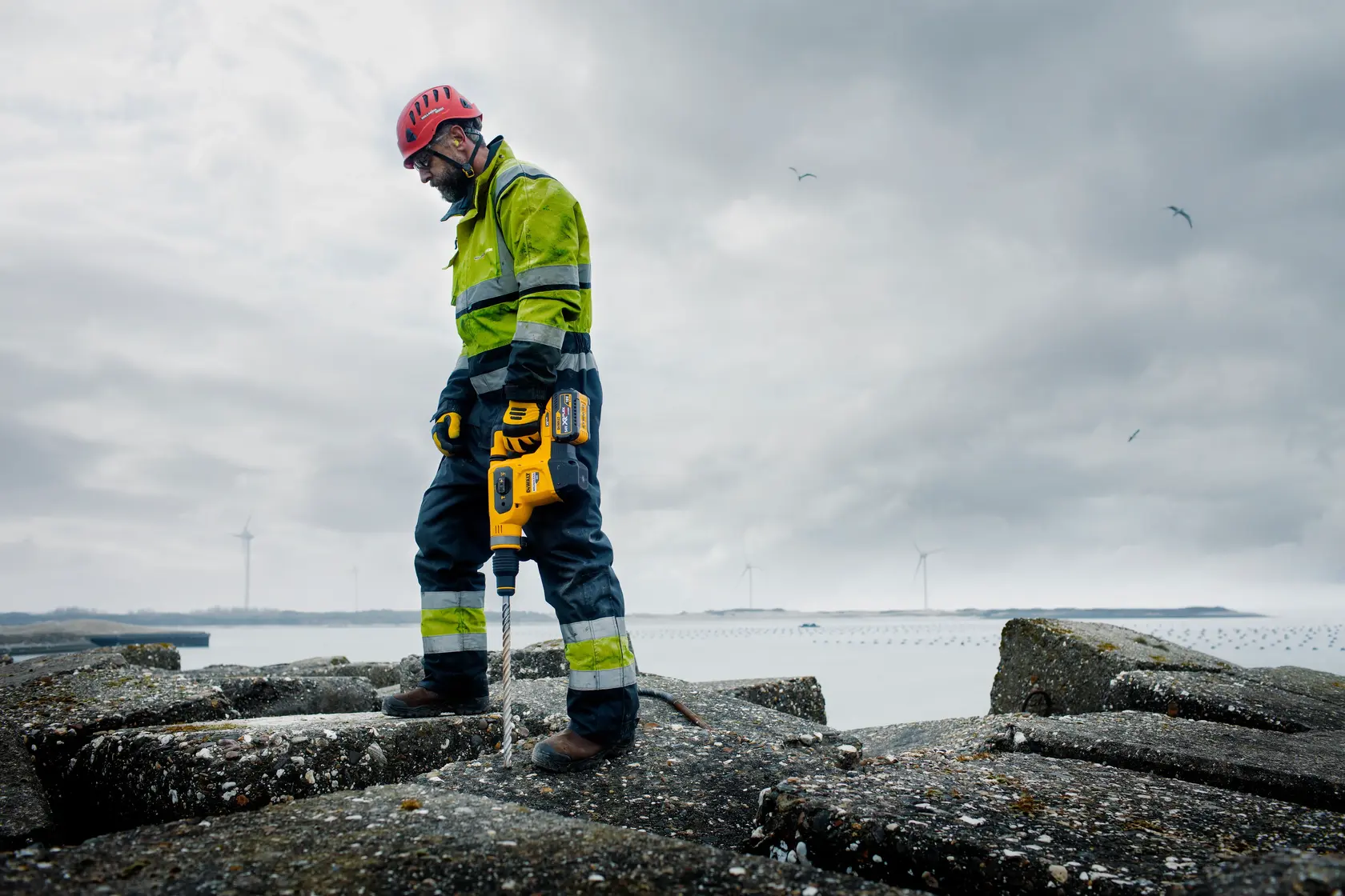A person wearing a high-visibility jacket, safety helmet, and gloves uses a yellow DEWALT power tool on a rocky surface outdoors. Wind turbines and water are visible in the background under a cloudy sky.