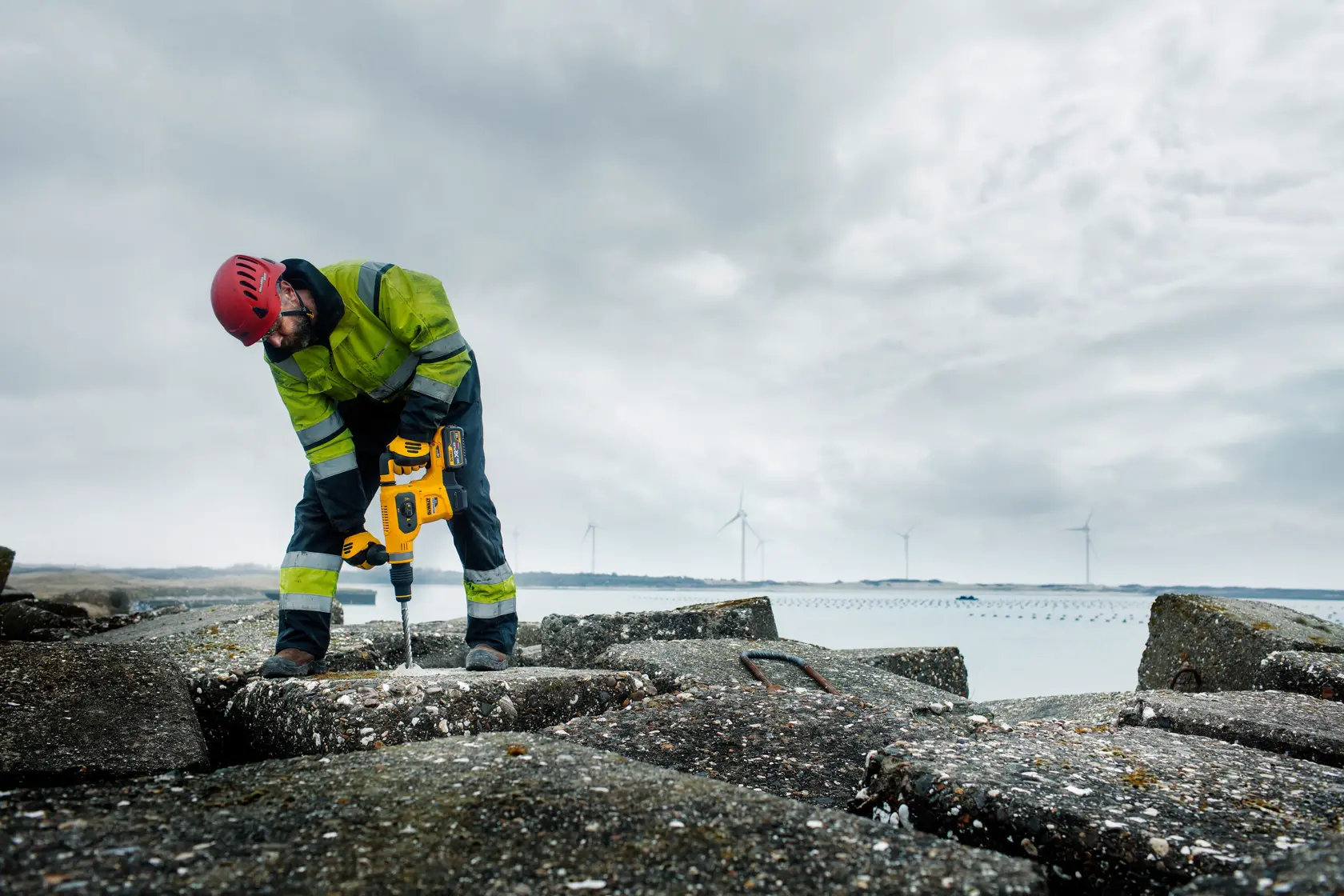 Worker using yellow DEWALT tool outdoors on concrete, wearing safety gear. Wind turbines in background.