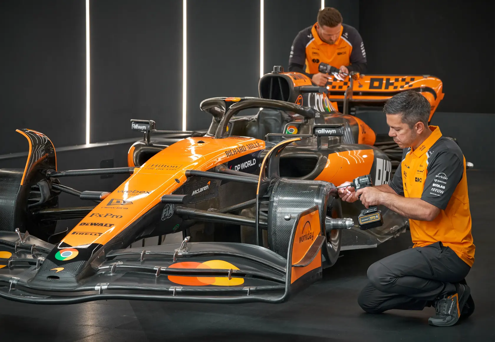 Two people work on an orange race car with tools in a modern garage. Faces are blurred for privacy.