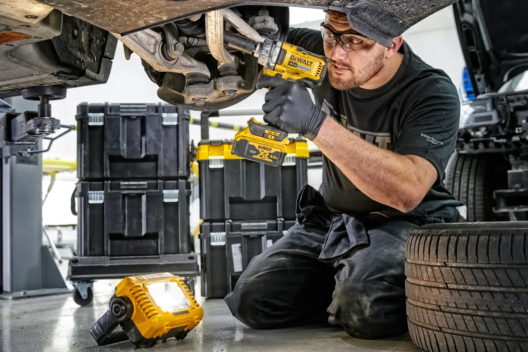 Person using DEWALT tools on car suspension, with DEWALT work light on floor and tool storage boxes nearby.