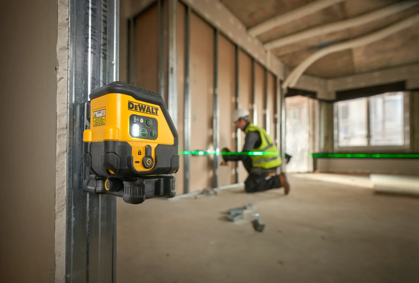 DEWALT laser level emits green line in construction site. Worker in high-visibility vest is installing wall panels.