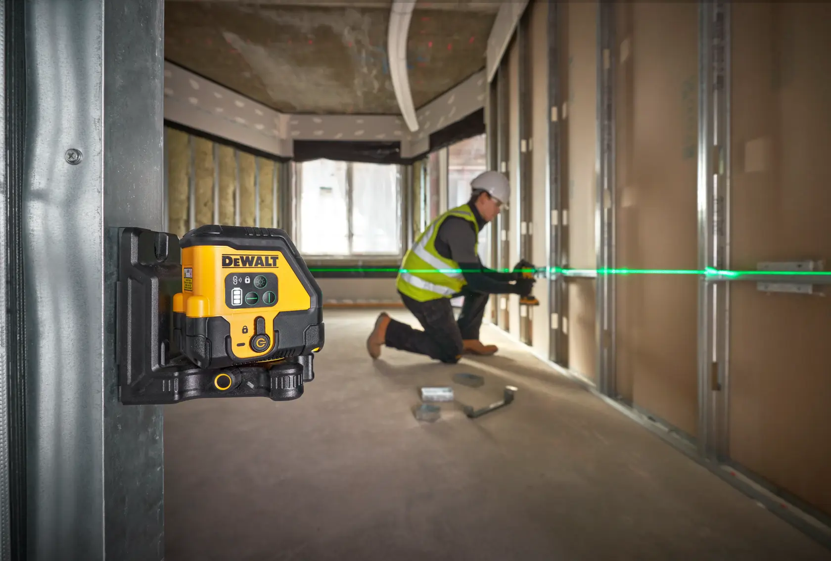 DEWALT laser level mounted on wall, projecting green line while worker aligns construction materials in an unfinished room.