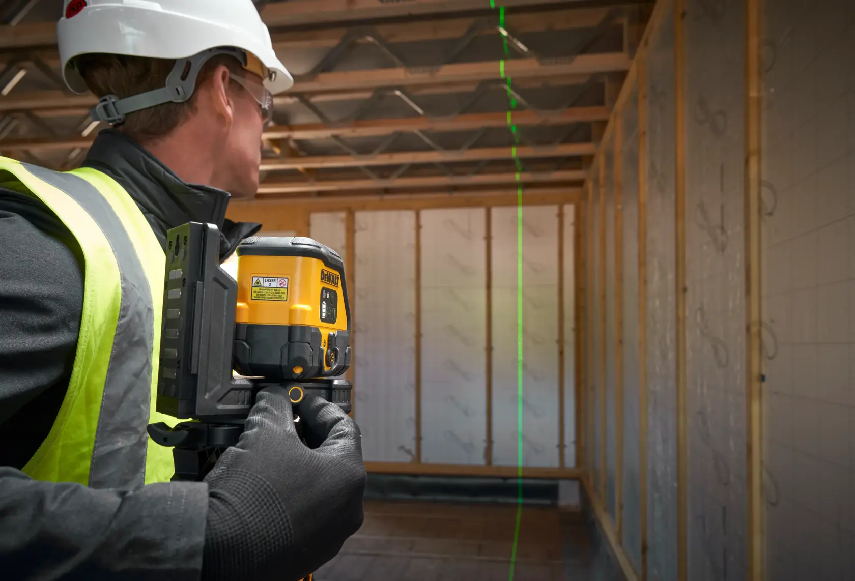 Worker uses DEWALT laser level in unfinished building, projecting a vertical green line for alignment.