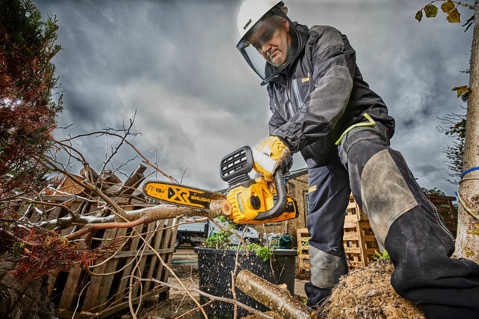 Person using a DEWALT cordless chainsaw to cut tree branches outdoors, wearing protective gear.