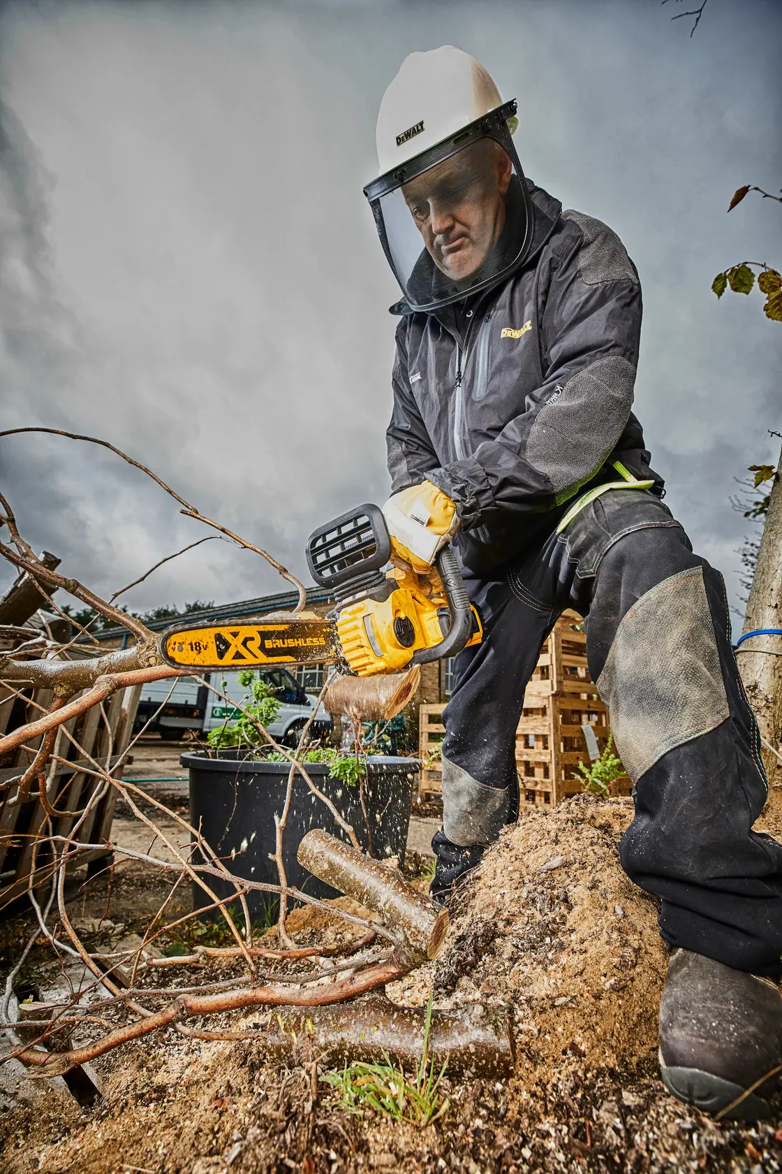 Person in safety gear uses a DEWALT chainsaw outdoors, cutting tree branches. Sawdust is visible.