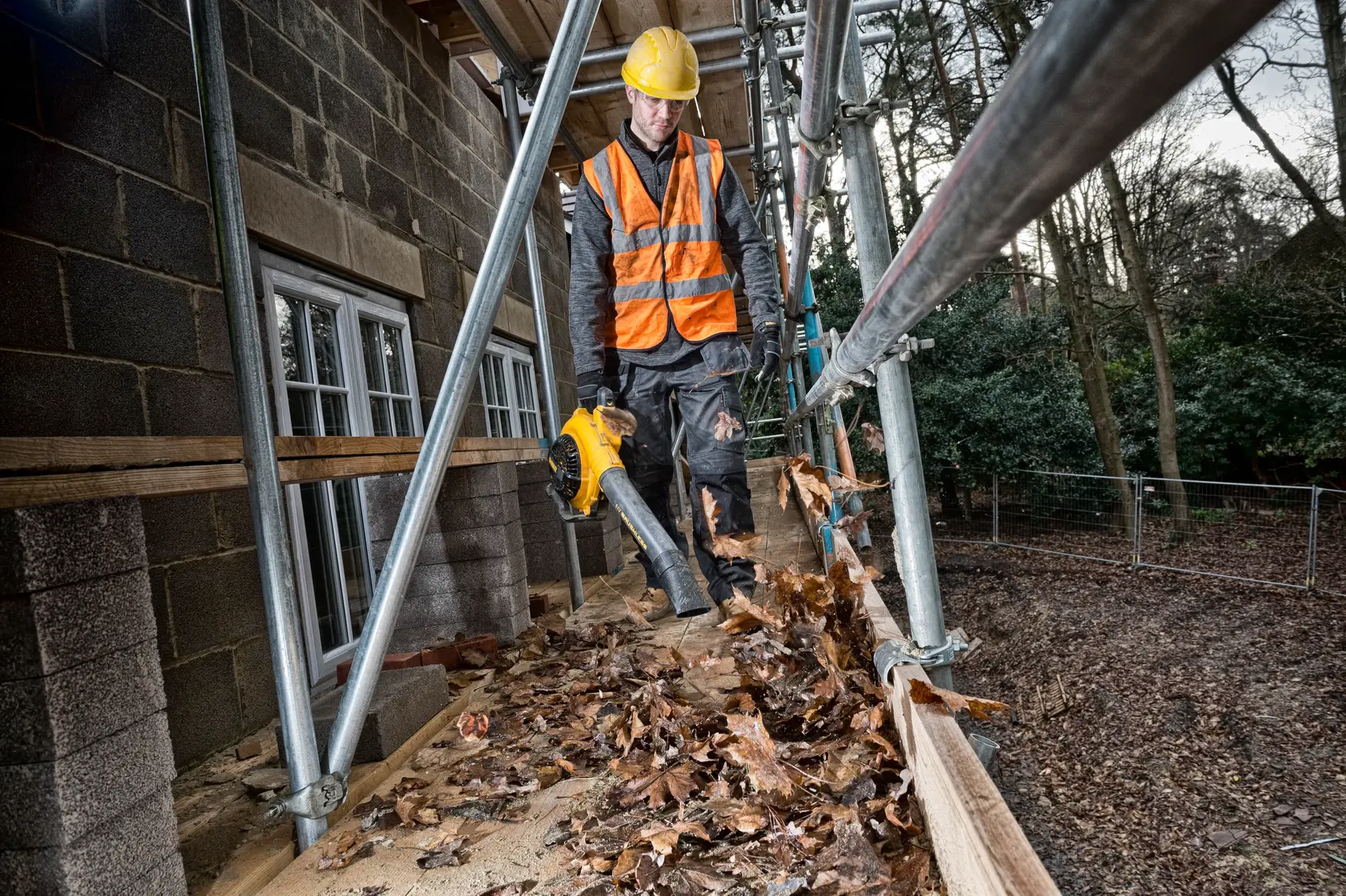 Worker uses a DEWALT leaf blower to clear leaves from scaffolding at a construction site.