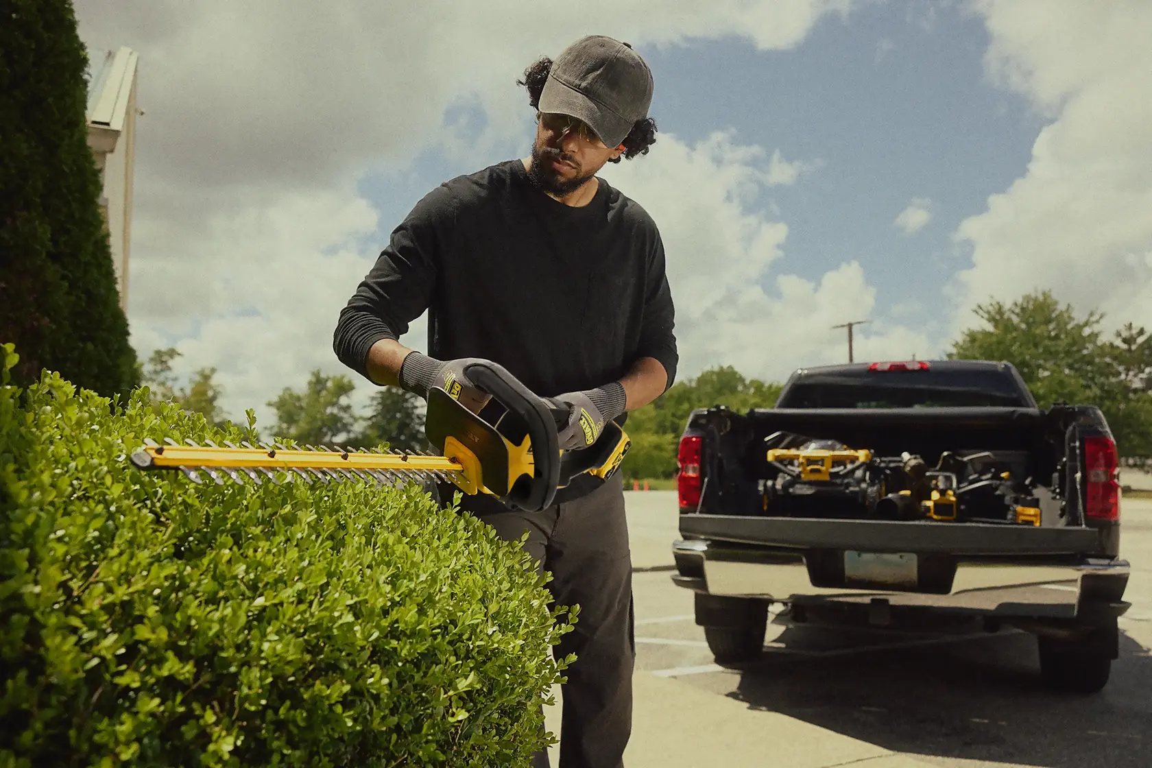 Person trims hedge with DEWALT cordless hedge trimmer, pickup truck with tools in background outdoors.