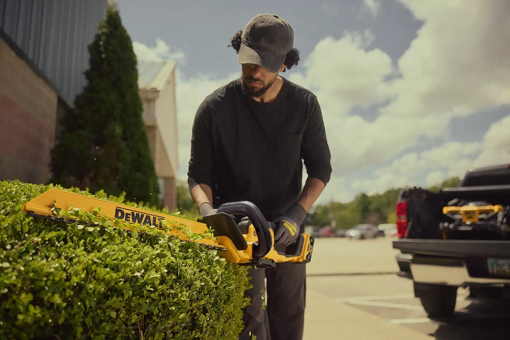 Person trims hedge with DEWALT cordless hedge trimmer outdoors near a parked truck on a sunny day.