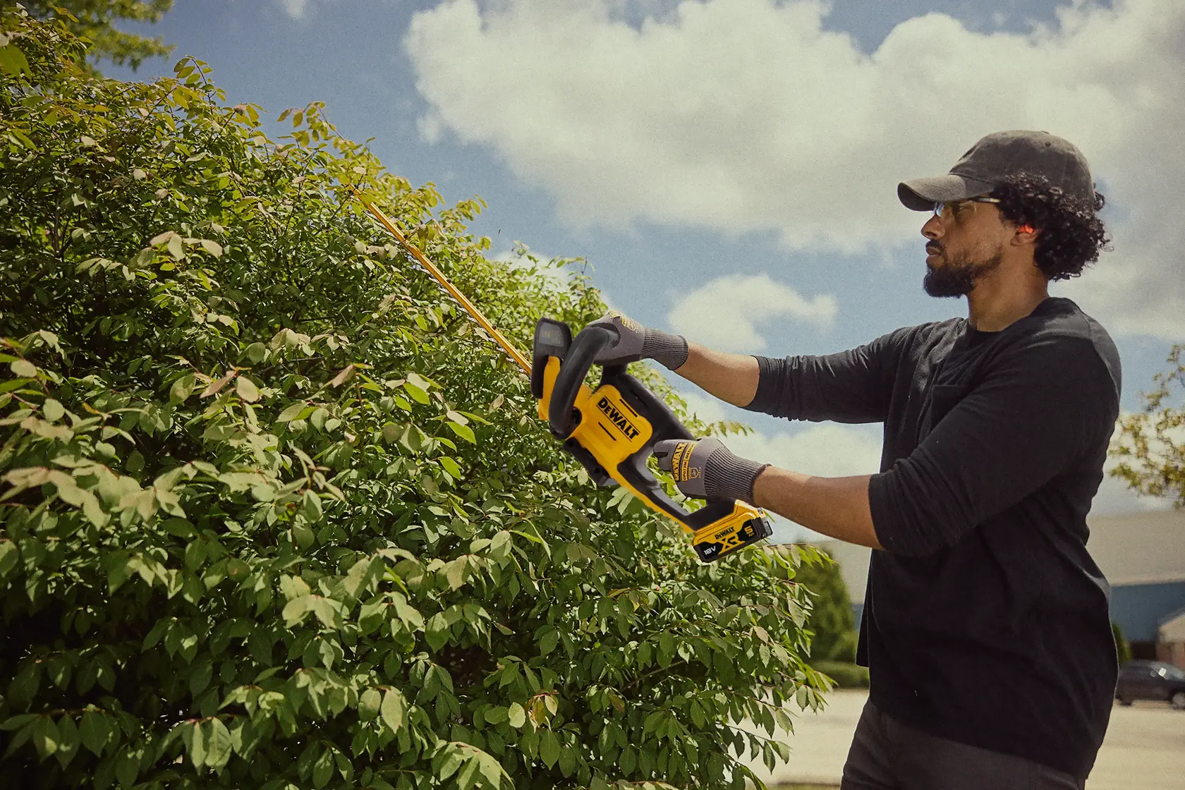 Person trims bushes using a DEWALT cordless hedge trimmer outdoors on a sunny day.