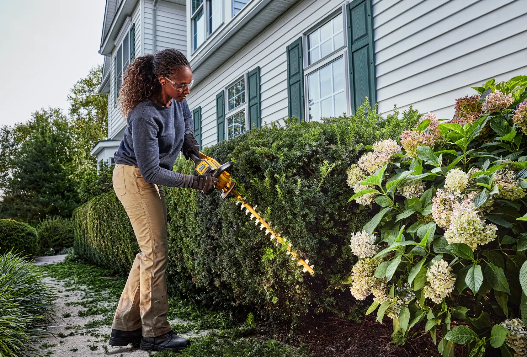 Person uses DEWALT cordless hedge trimmer to trim bushes in a garden outside a house.