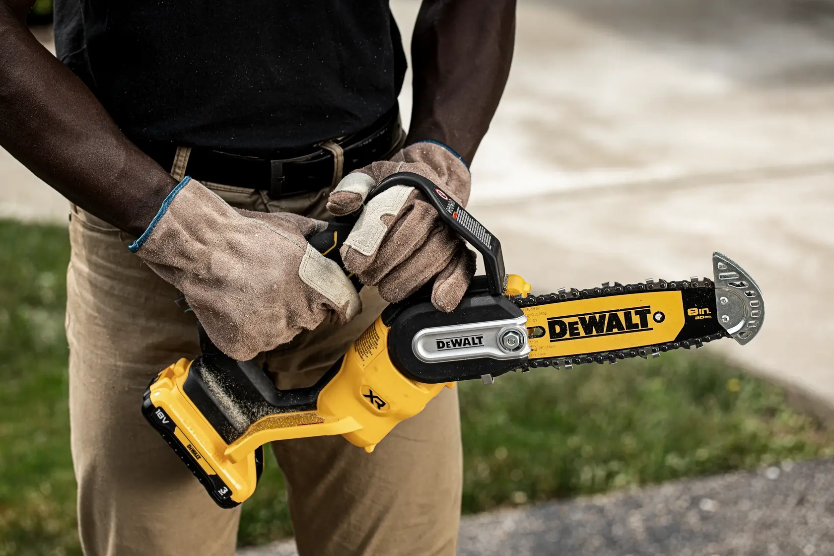 Person wearing gloves holding a DEWALT cordless chainsaw outdoors close to their body.