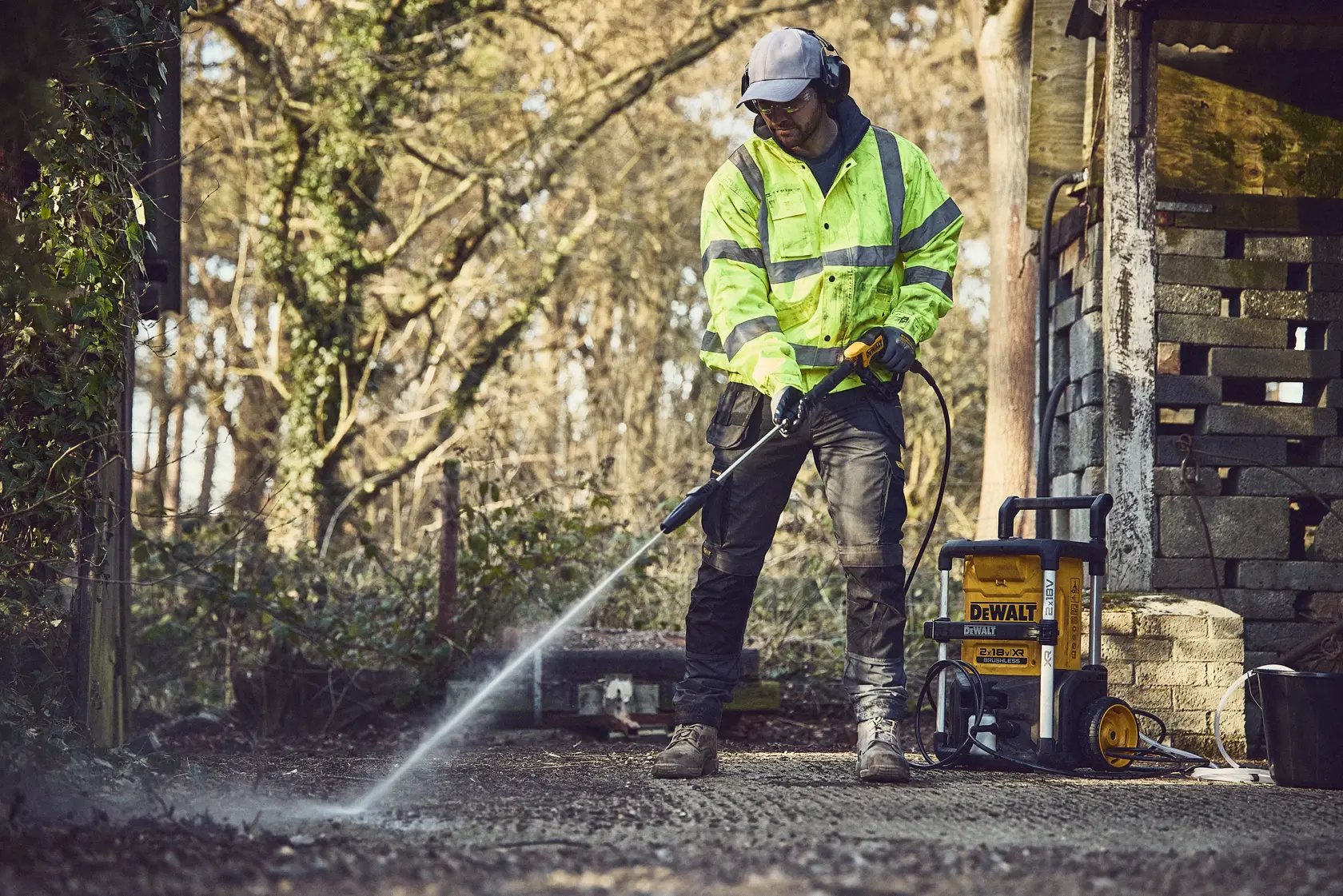Person uses a DEWALT pressure washer outdoors on a paved surface beside a shed, wearing safety gear.