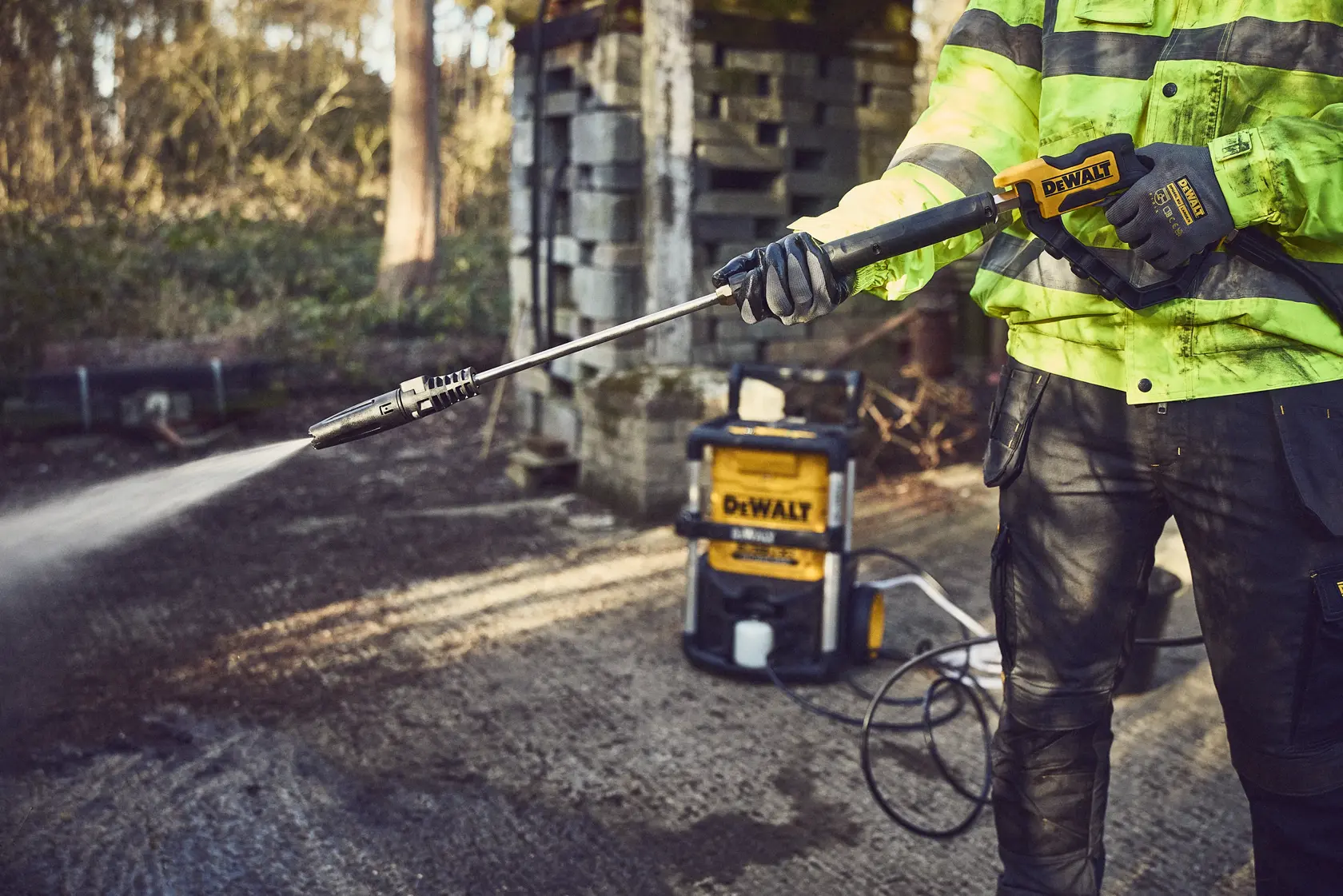 A person uses a DEWALT cordless pressure washer outdoors, spraying water on the ground.