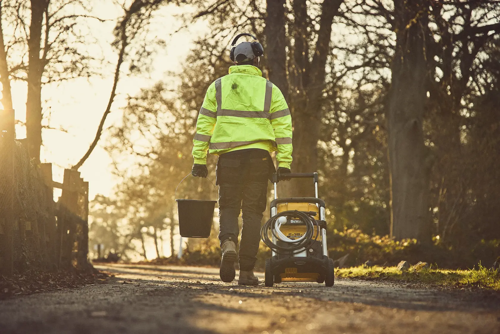 Person in safety gear pulls a DEWALT power washer and carries a bucket on a rural path at sunset.