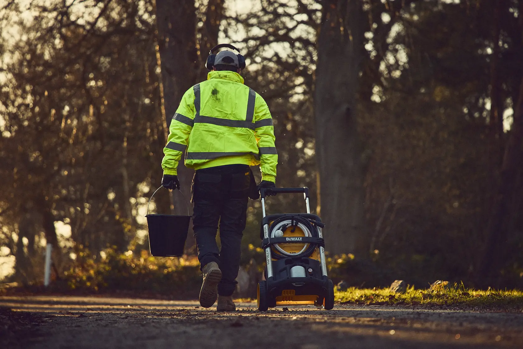 Worker in high-vis jacket pulls DEWALT pressure washer and carries bucket outdoors on path.