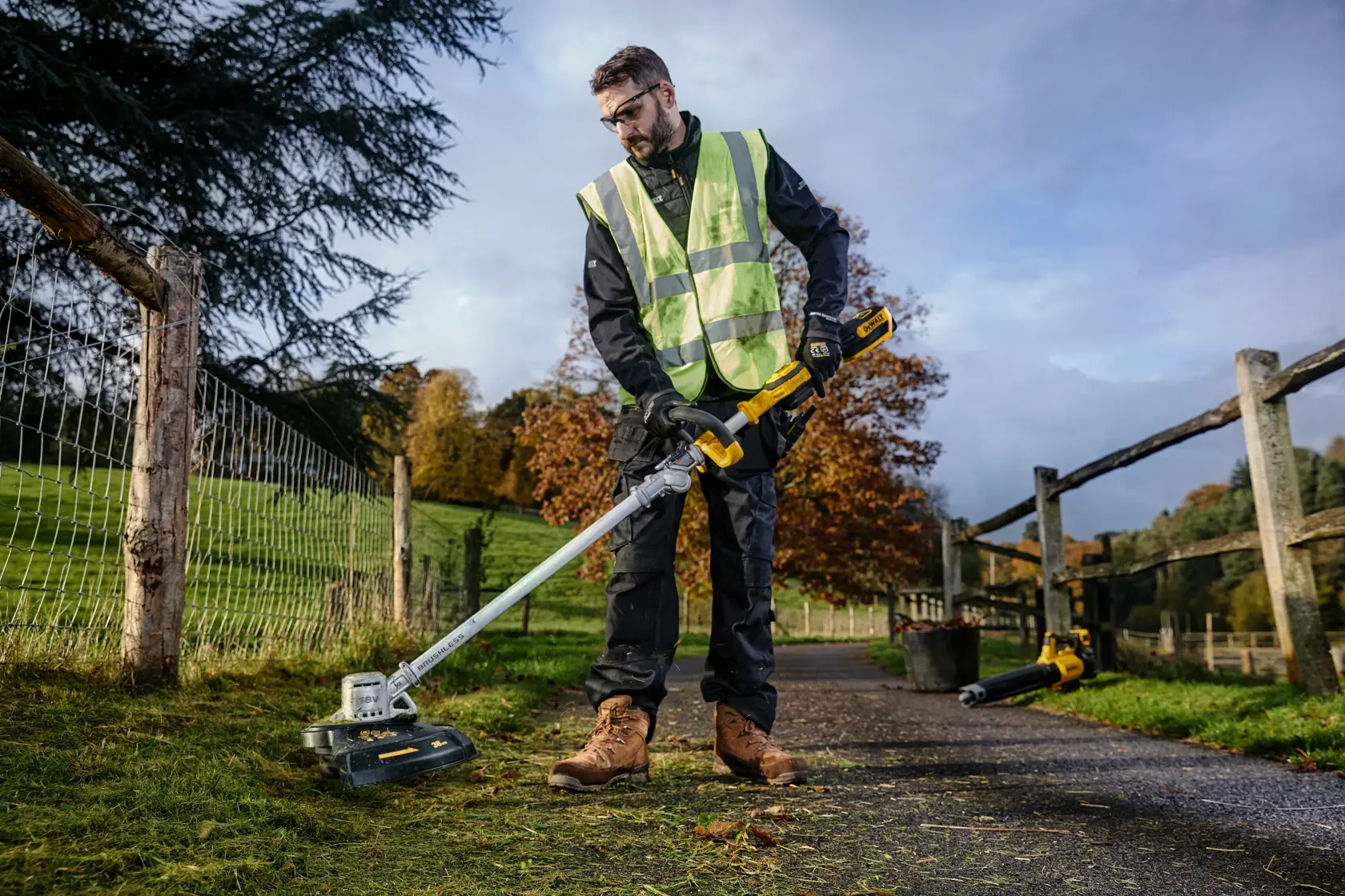 Worker uses a DEWALT DCMST561N string trimmer on grass near a fence in an outdoor setting.