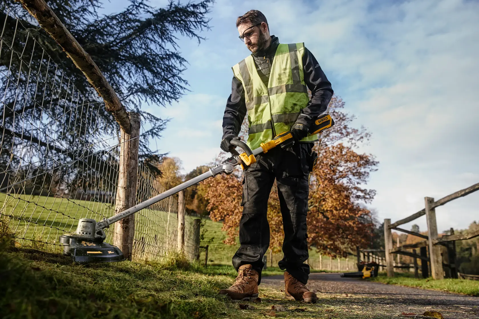 Person uses DEWALT DCMST561N grass trimmer outdoors near a fence on a sunny day.