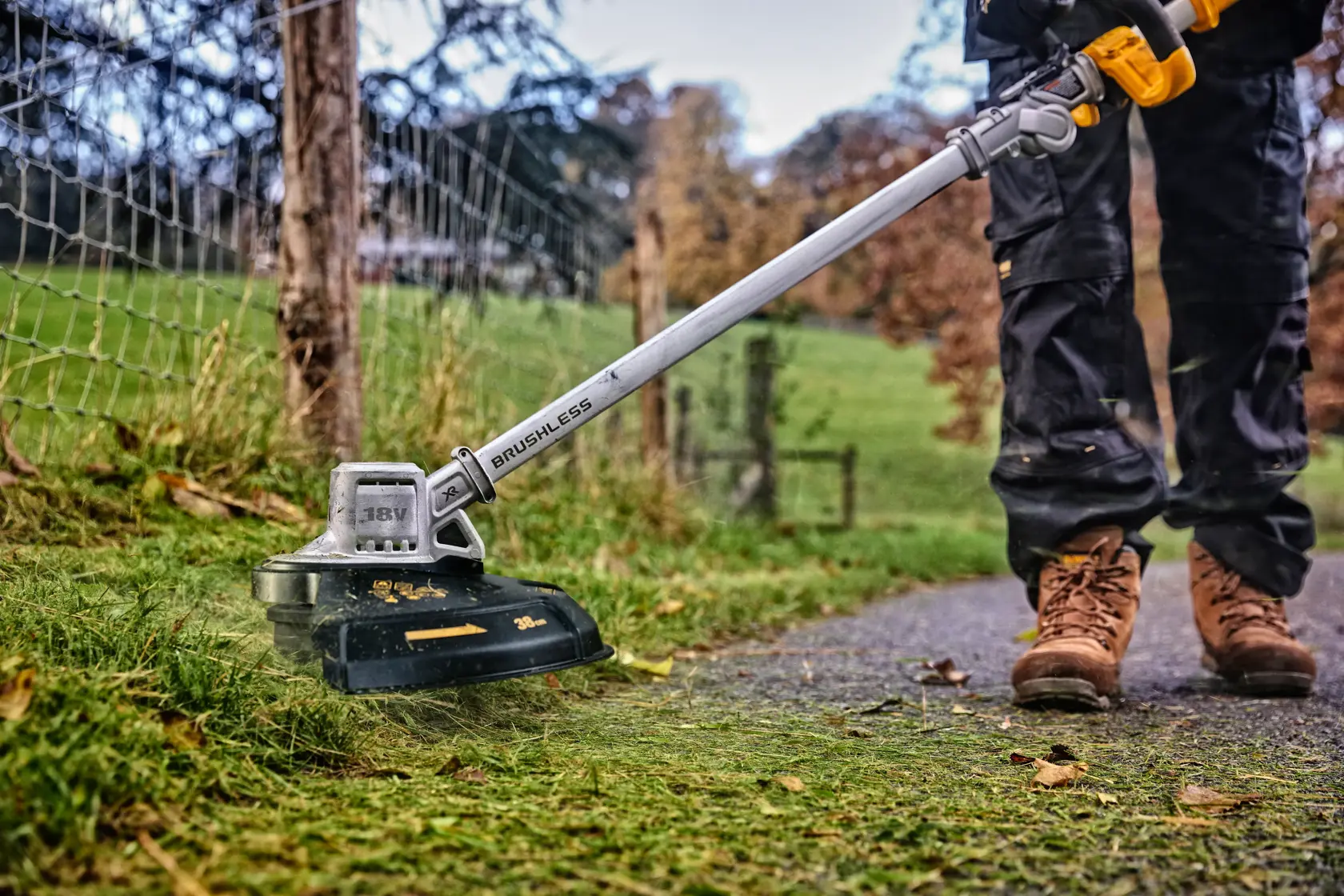 DEWALT cordless brushless grass trimmer used outdoors near a fence, operator in work boots and pants visible.
