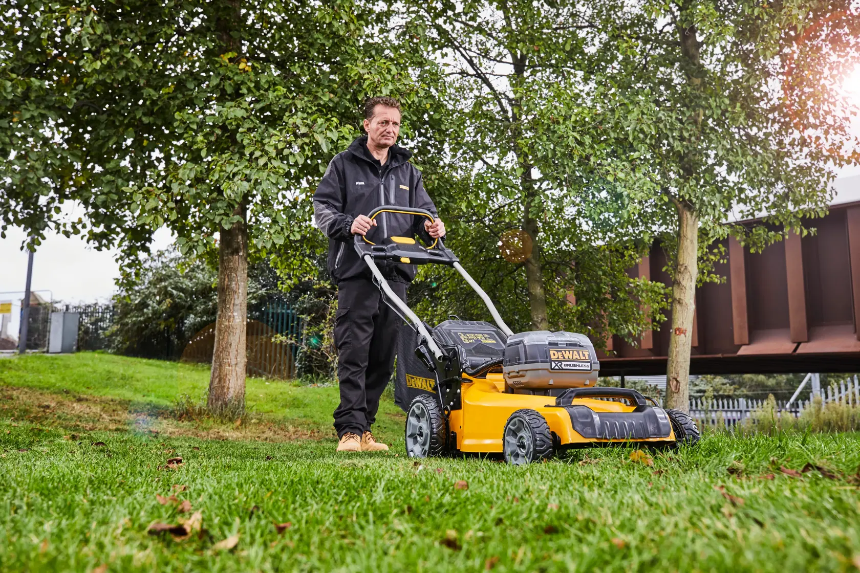 Person using a DEWALT DCMW564P2 lawn mower outdoors on grass near trees.