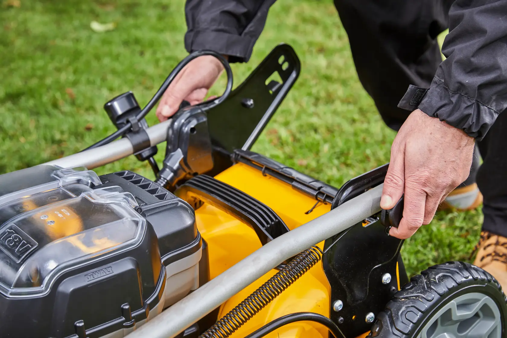 Close-up of hands adjusting the handle on a DEWALT cordless lawn mower outdoors on grass.