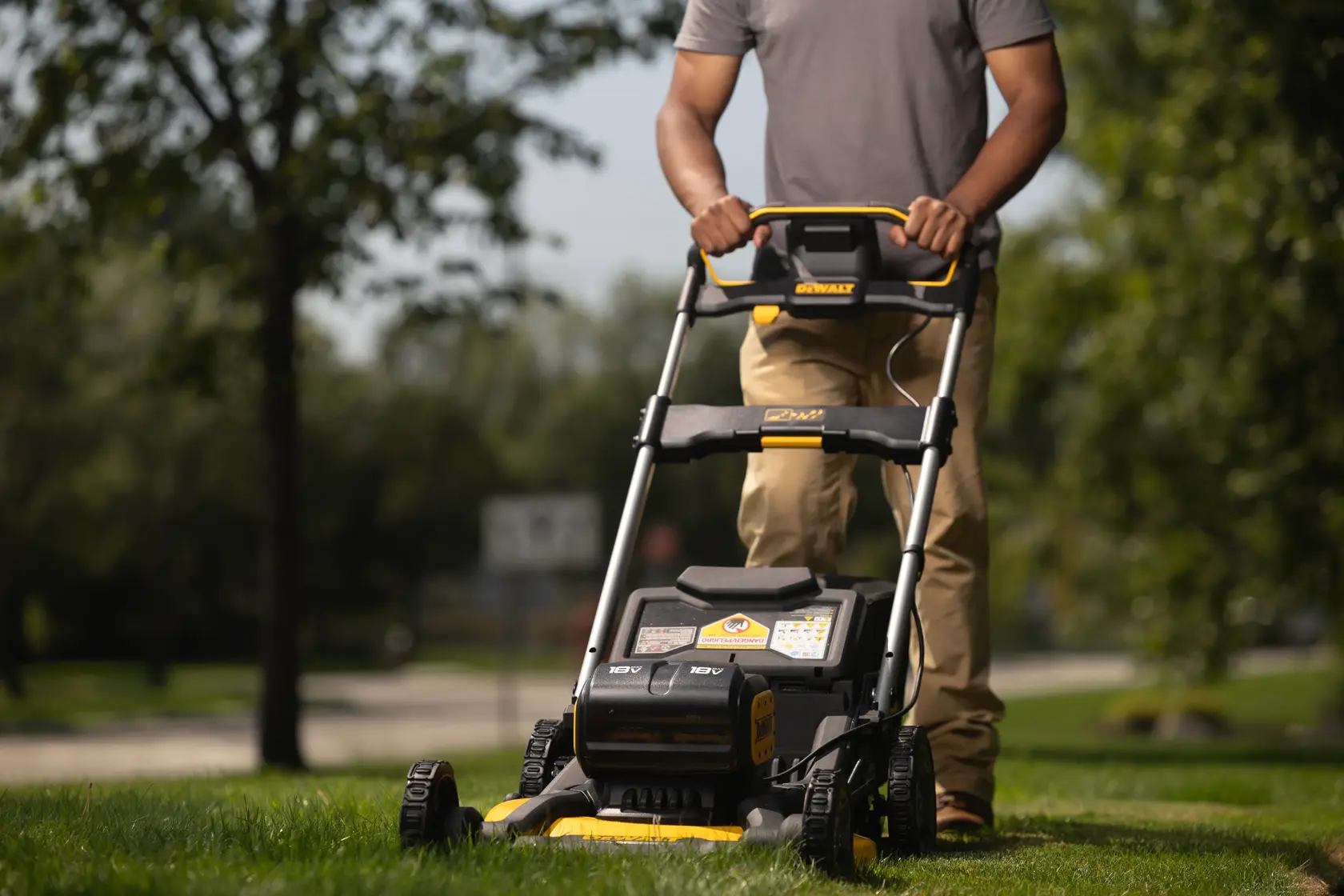 A person is pushing a DEWALT cordless electric lawn mower on green grass outdoors, with trees and a road visible in the background.