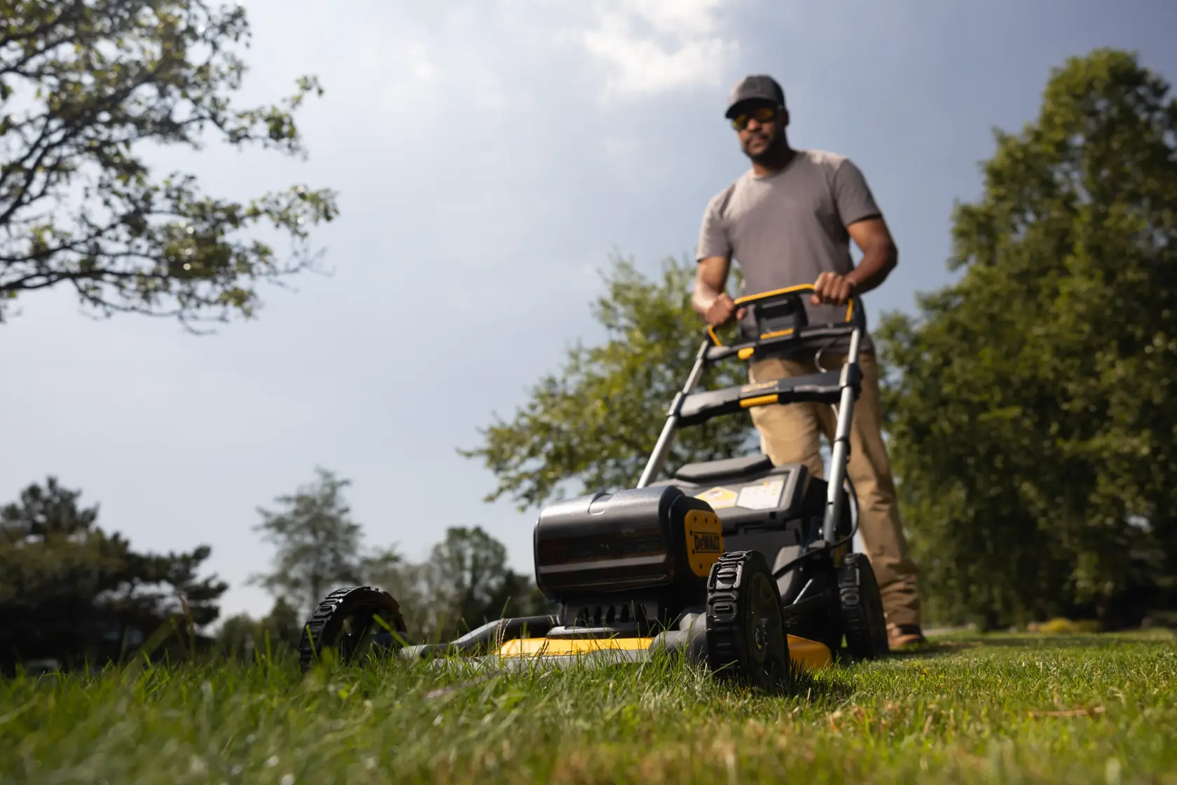 A person operates a DEWALT cordless lawn mower, model DCMWP134W2, outdoors on a grassy lawn with trees in the background. The person's face is blurred.