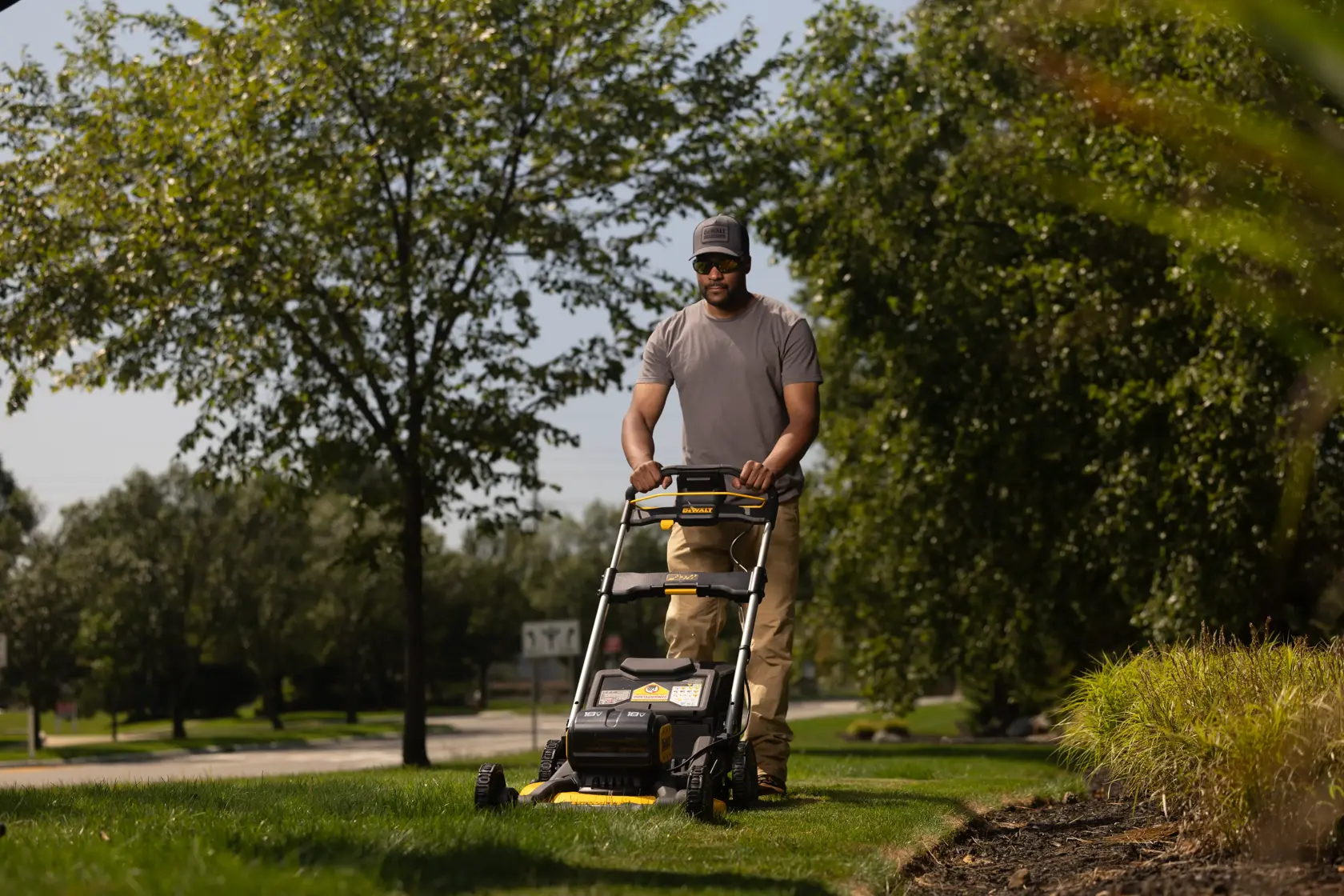 A person using a DEWALT cordless lawn mower (model DCMWP134W2) to cut grass in a garden or outdoor area, with trees and plants in the background.