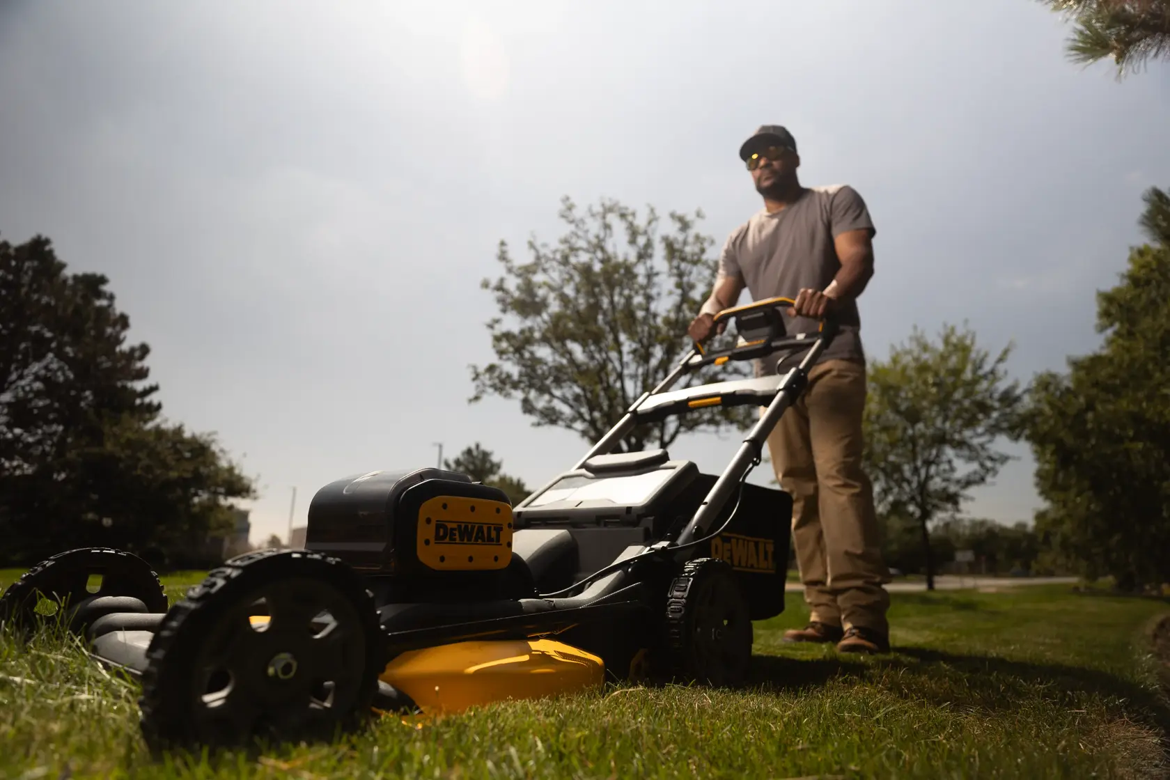 A person is pushing a DEWALT lawn mower (model DCMWP134W2) on a grassy area outdoors. The mower is black and yellow, and the person's face is blurred for privacy. There are trees and a cloudy sky in the background.