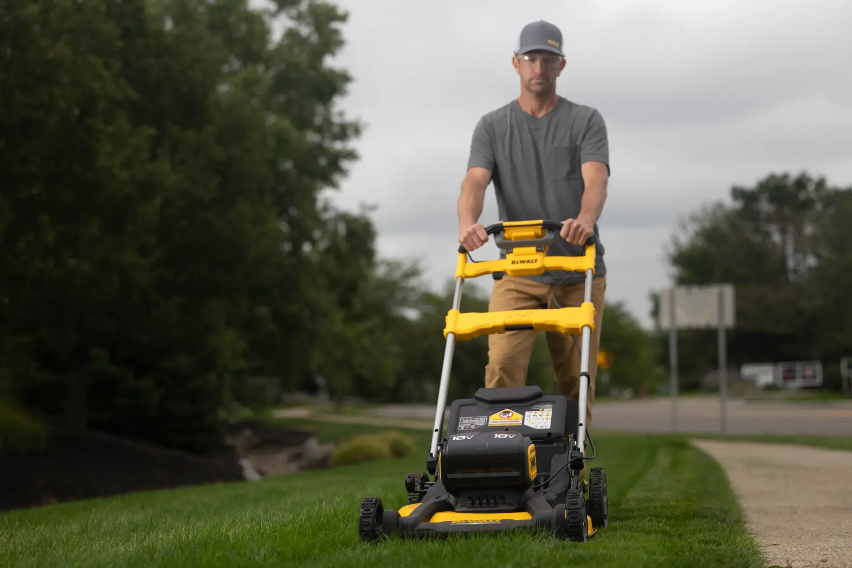 A person is pushing a yellow and black DEWALT lawn mower on green grass in a residential outdoor setting. The person's face