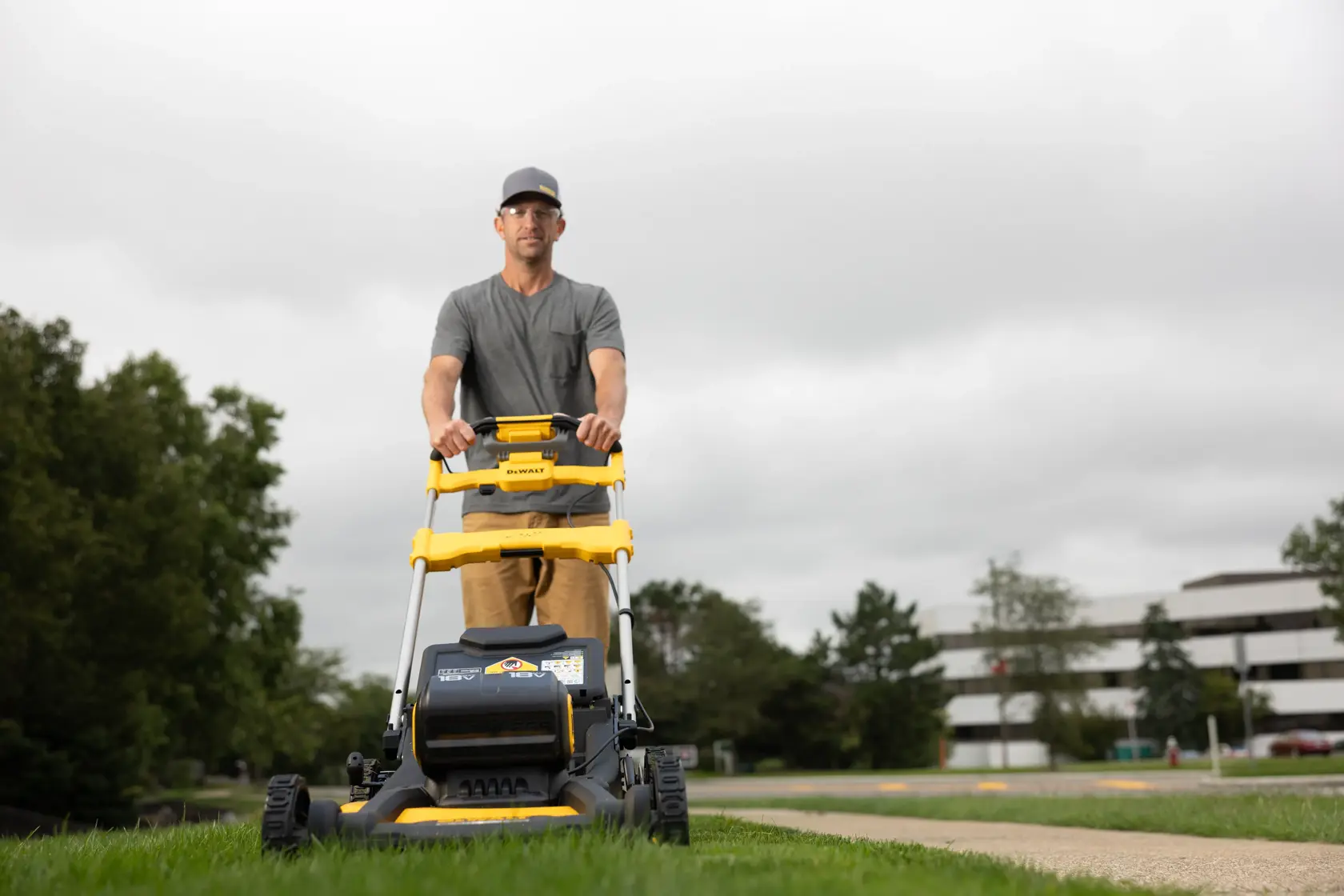 A person is pushing a DEWALT lawn mower on a grassy area outdoors. The image shows the mower from the front, and the