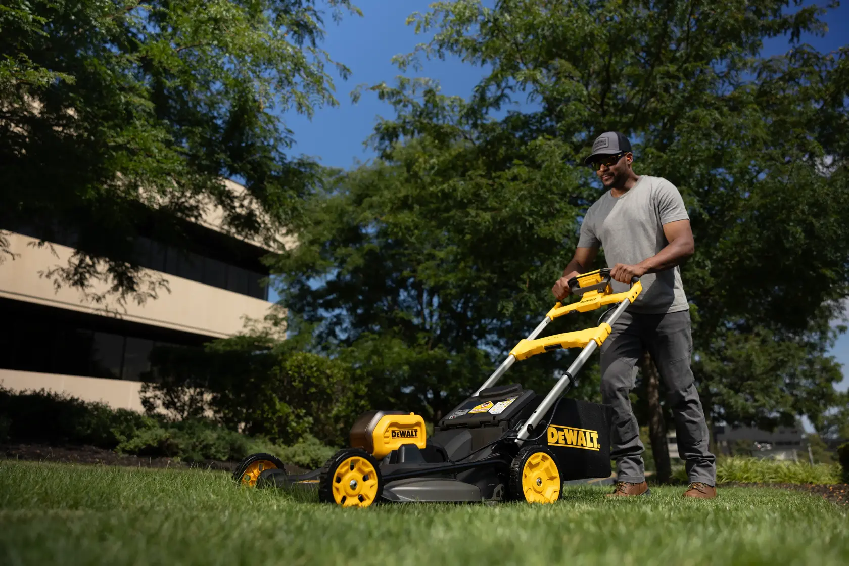 Person uses a DEWALT lawn mower outdoors on grass with buildings and trees in the background.