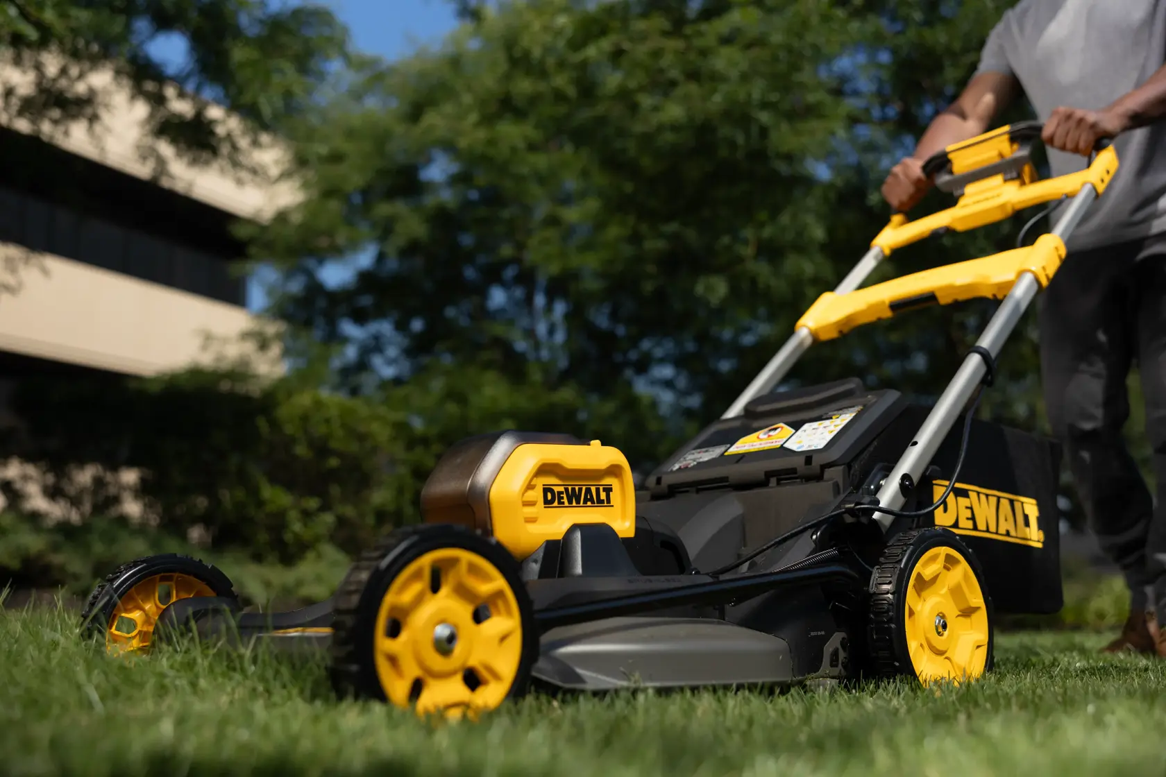 A DEWALT lawn mower is being pushed on grass outdoors, with trees and a building in the background.