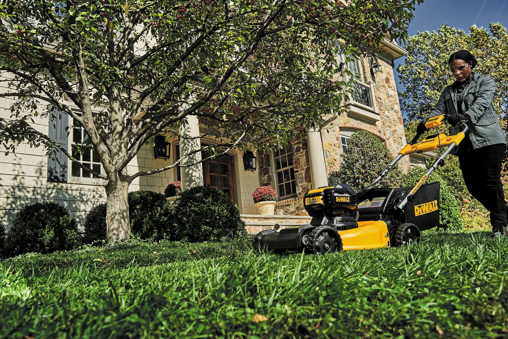 Person using a DEWALT lawn mower on grass in front of a house with trees and bushes on a sunny day.