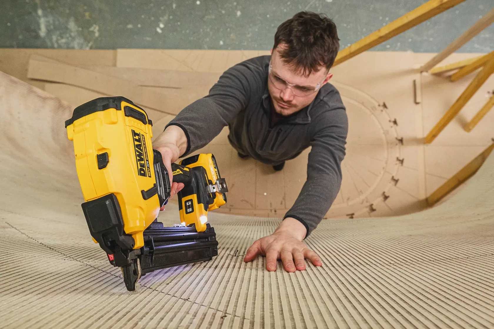 Person using a DEWALT nailer on curved wooden surface, wearing safety glasses and gray top.