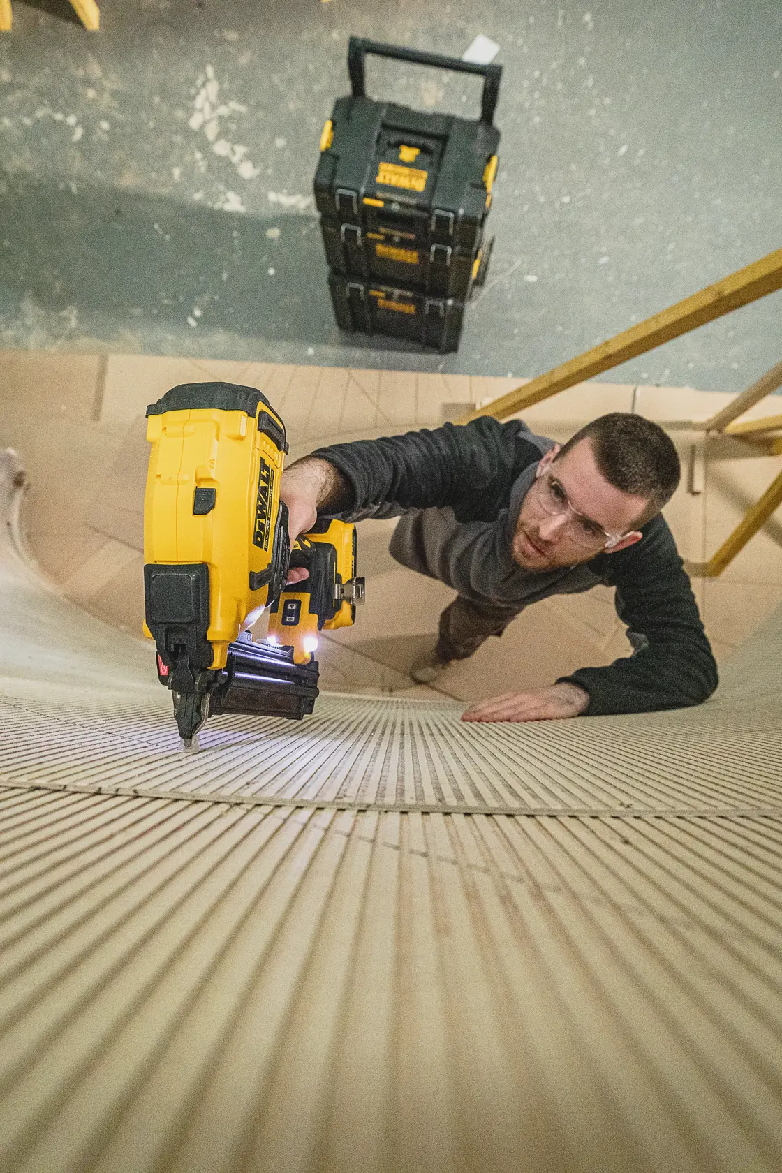Person using a DEWALT nailer tool on wood surface, with DEWALT storage boxes visible in the background.