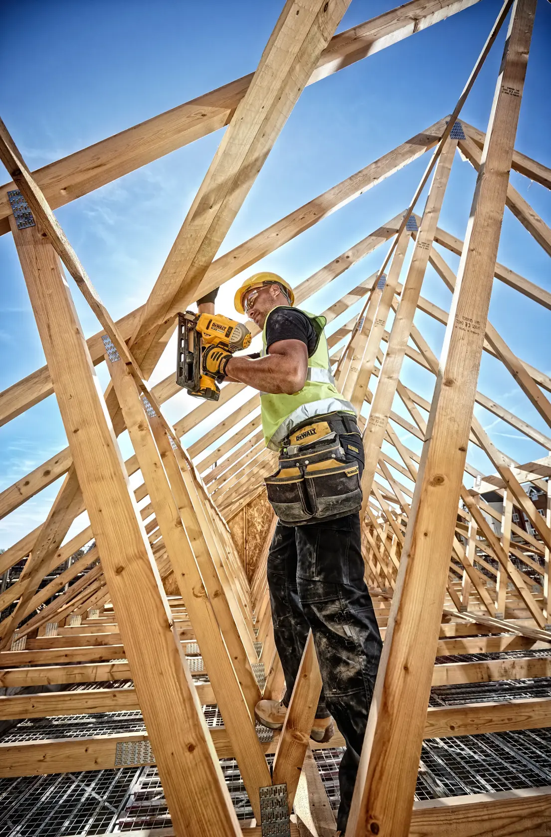 Worker uses a DEWALT nailer for wood construction on a roof, wearing safety gear and tool belt.