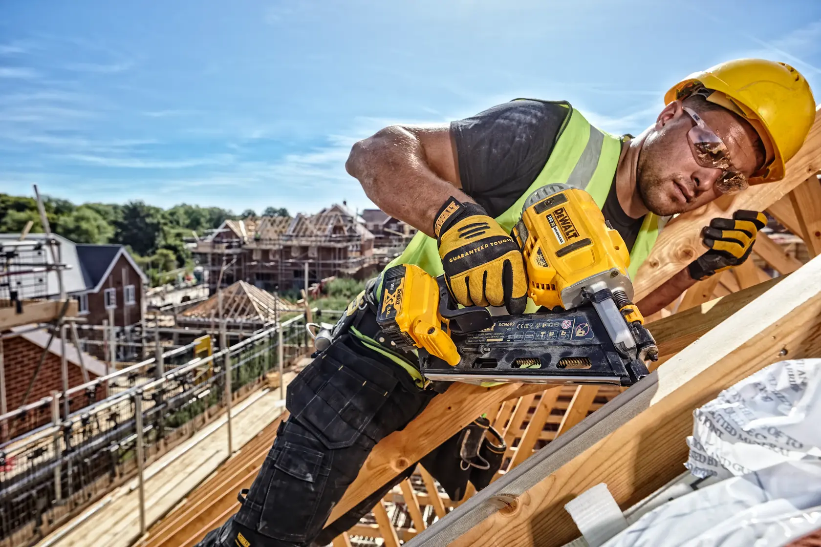 Worker uses a DEWALT cordless nailer DCN692N on a wooden roof frame at a construction site.