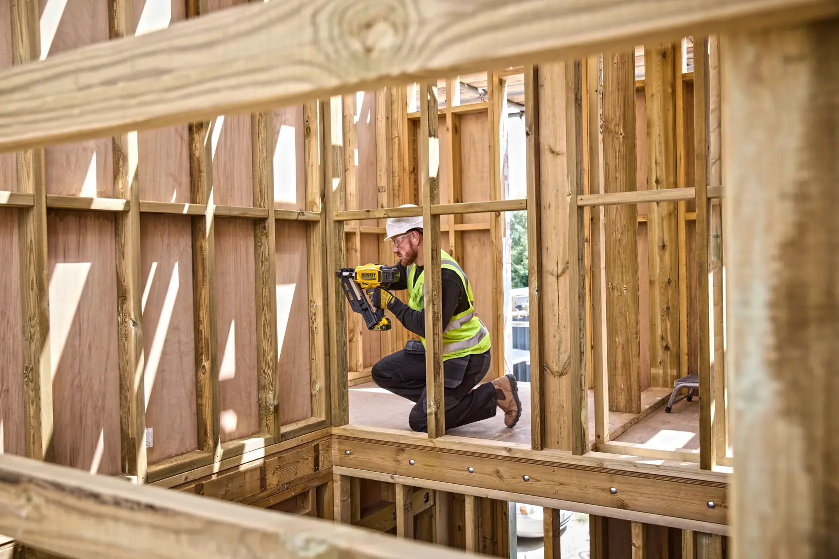 Worker uses a DEWALT tool to fasten wood frames at a construction site, wearing safety gear and kneeling.