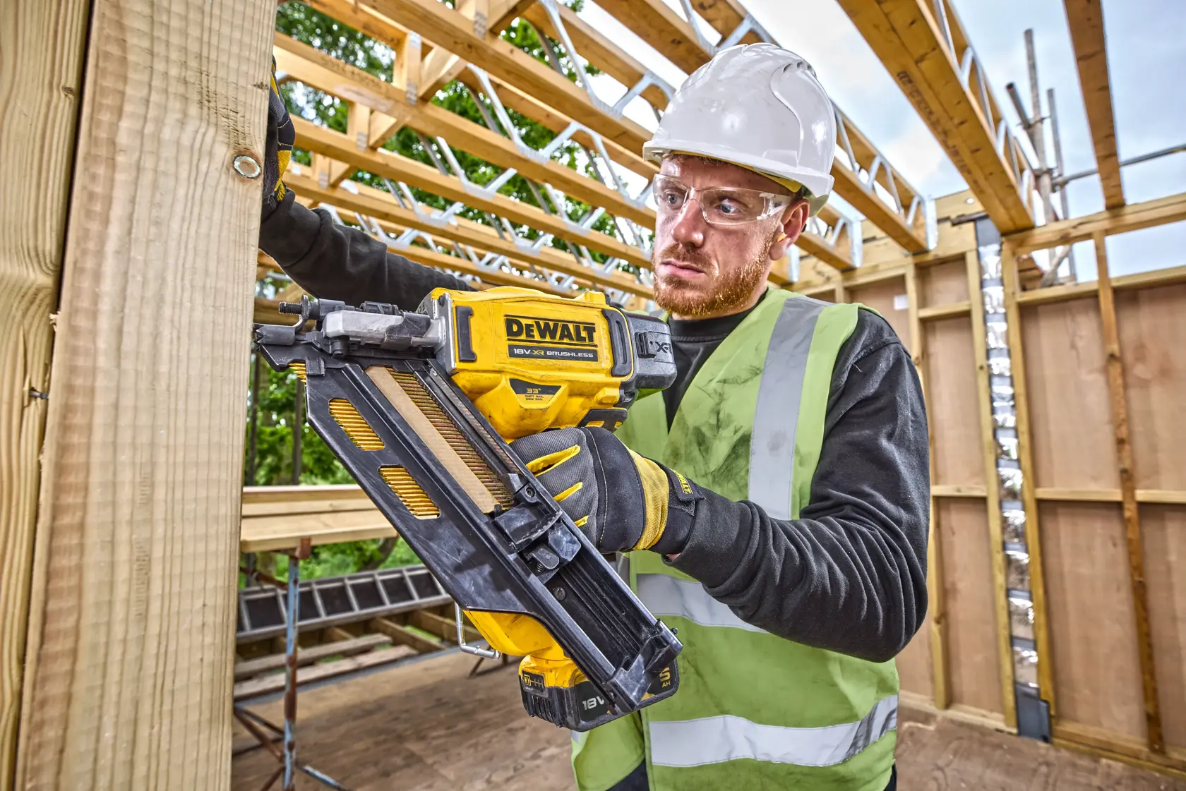 Person uses a DEWALT nailer to secure wood at a construction site, wearing a hard hat and high-visibility vest.